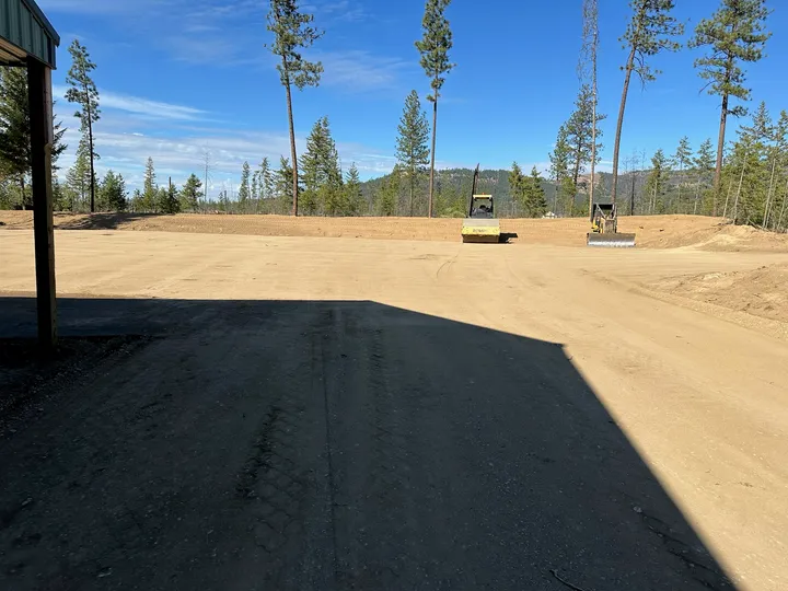Wide view of a graded building pad with compacted gravel and pine forest behind it