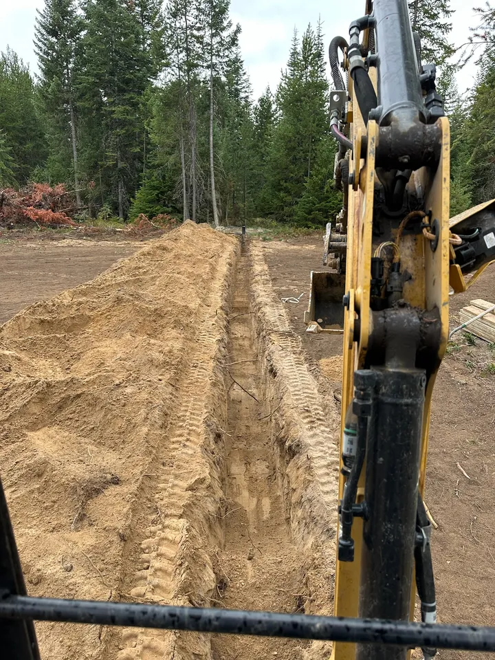 Open utility trench seen from the excavator cab, running across a lot toward a house